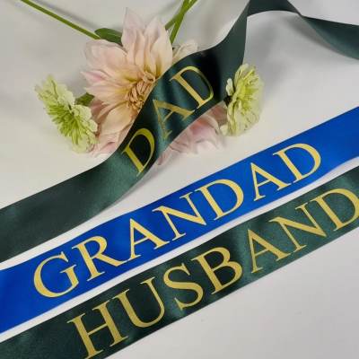 Memorial ribbons reading Dad, Grandad, and Husband with pale flowers in the background