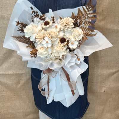 Handheld bouquet of cream roses and white blooms with brown foliage, wrapped in white paper.