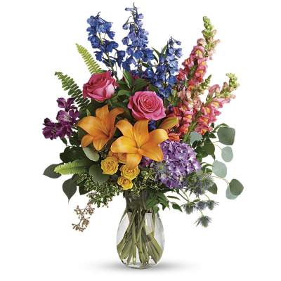 Tall mixed bouquet of orange lilies, pink roses, and assorted bright flowers in a clear glass vase