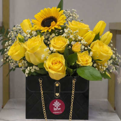 Yellow roses and a sunflower arranged in a black gift box with a gold chain.