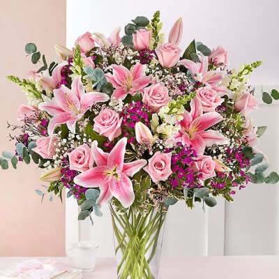Tall glass vase filled with pink lilies, roses, and white accent flowers.