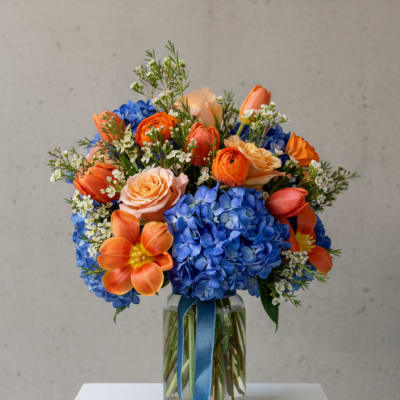 Bouquet of blue hydrangeas, orange tulips, and peach roses in a glass vase