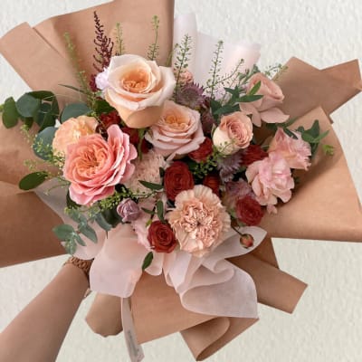 Hand-tied bouquet of peach and pink roses with carnations in brown paper