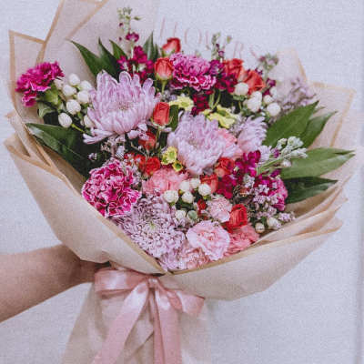 Handheld bouquet of pink and red flowers wrapped in beige paper with a pink ribbon