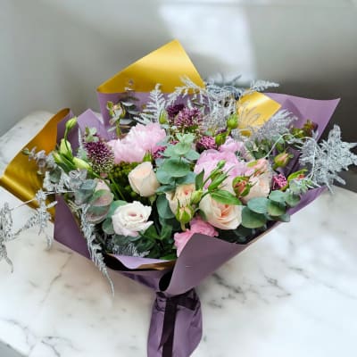 Hand-tied bouquet of pink and cream flowers with purple accents wrapped in purple and gold paper