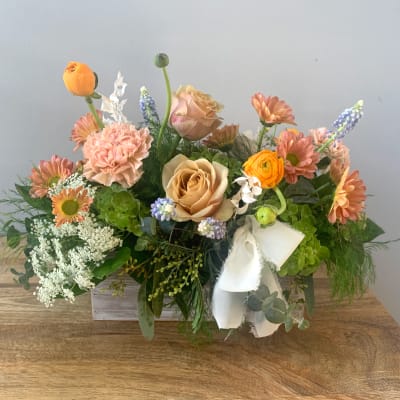 Low arrangement of peach roses, carnations, and orange blooms in a white wooden box with a fabric bow