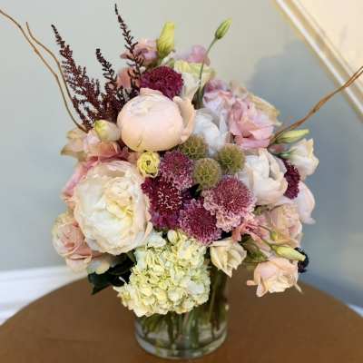 Pastel bouquet of roses, peonies, hydrangea, and purple accent flowers in a glass vase