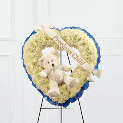 Heart-shaped white flower wreath with a teddy bear and ribbon on a stand