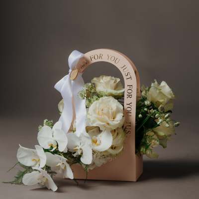 Cream roses and white orchids arranged in a blush gift box with a ribbon handle.