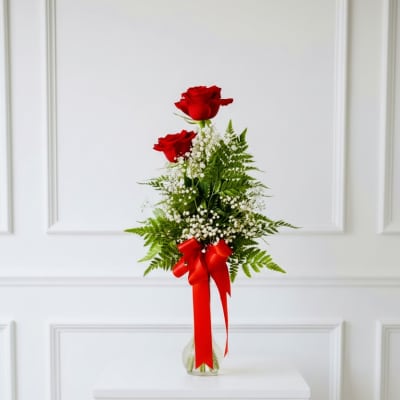Two red roses in a clear vase with baby's breath and a red ribbon
