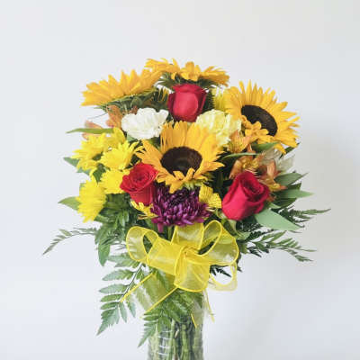 Bouquet of sunflowers, red roses, and mixed blooms in a glass vase with a yellow bow