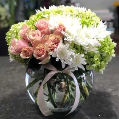 Round arrangement of pink roses, white mums, and green hydrangeas in a clear bowl vase with ribbon