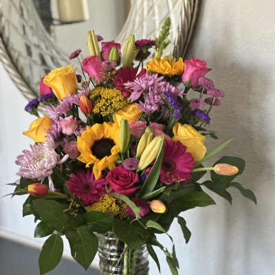 Tall mixed bouquet of sunflowers, roses, lilies, and other bright blooms in a clear glass vase
