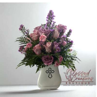 Pink and lavender roses with purple filler flowers in a white vase with a cross