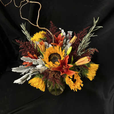Sunflowers and orange lilies arranged in a glass vase with curly branches