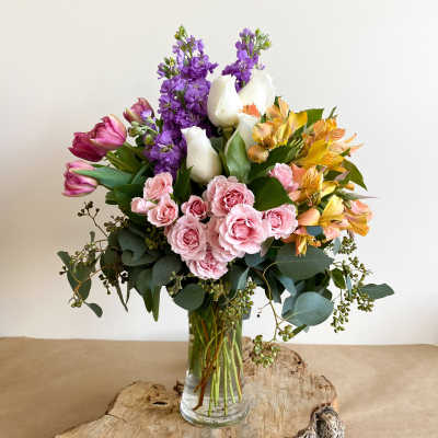 Mixed bouquet of pink roses, purple stock, white tulips, and yellow alstroemeria in a glass vase
