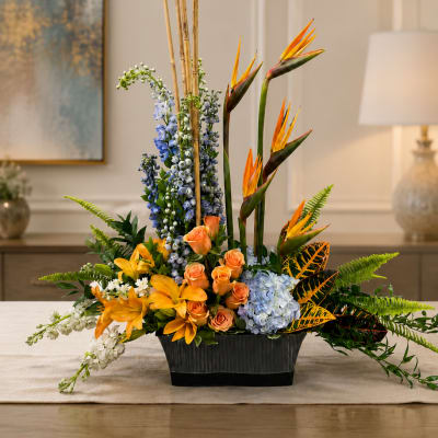 Tropical floral arrangement with orange birds of paradise, roses, lilies, and blue blooms in a black vase
