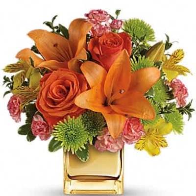 Bright orange lilies and roses with mixed blooms in a gold cube vase