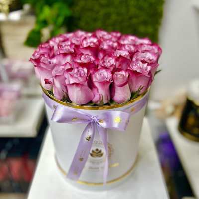 Round white box filled with tightly packed pink roses, finished with a lavender ribbon bow
