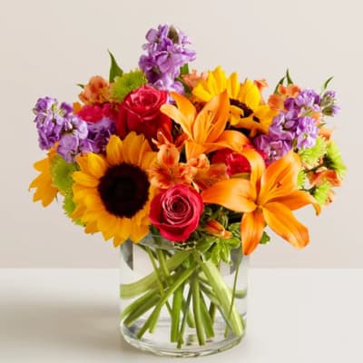 Mixed bouquet of sunflowers, roses, lilies, and purple flowers in a glass vase