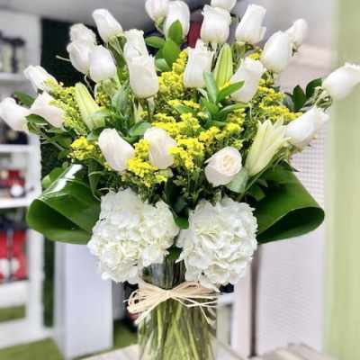 Tall arrangement of white roses, lilies, and hydrangeas with yellow accents in a clear glass vase