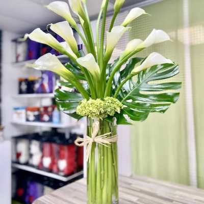 Tall arrangement of white calla lilies with large glossy leaves in a clear glass vase.