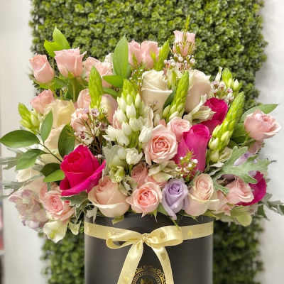 Round hatbox of mixed pink, cream, and purple roses with white accent flowers and a yellow ribbon bow.