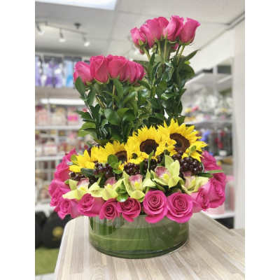 Low glass vase of bright pink roses, yellow sunflowers, green orchids, and grapes arranged in tiers.
