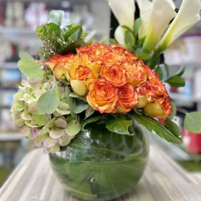 Low round arrangement of orange roses, green hydrangea, and white calla lilies in a glass bowl vase