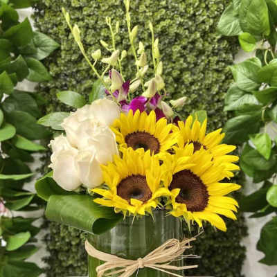 Arrangement of sunflowers, white roses and orchids in a glass vase wrapped with a green leaf and raffia bow