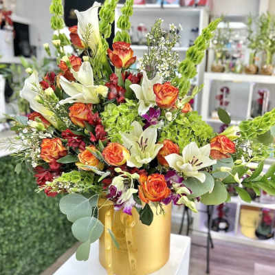 Tall arrangement of orange roses and white lilies in a gold hatbox-style container.