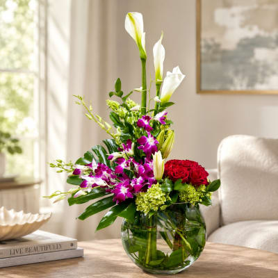 Tall floral arrangement with white calla lilies, purple orchids, and red roses in a glass vase.