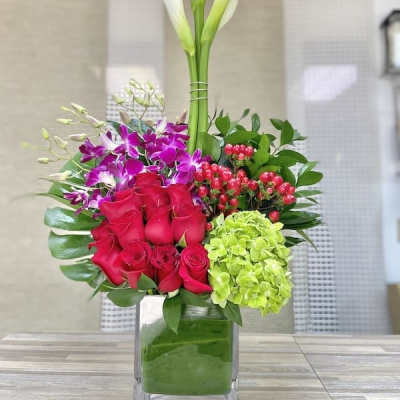 Tall modern arrangement with white calla lilies, red roses, purple orchids, and green hydrangea in a glass cube vase.