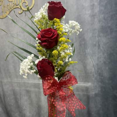 Three red roses in a glass vase with white and yellow filler flowers and a red ribbon