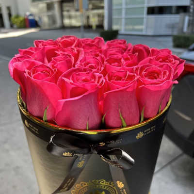 Bright pink roses arranged in a round black hatbox with gold accents and ribbon