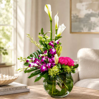 Tall floral arrangement with white calla lilies, purple orchids, and pink roses in a glass vase.