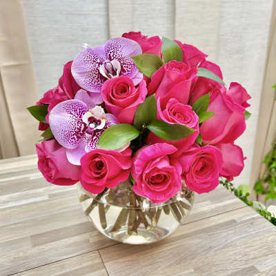Round arrangement of hot pink roses with two spotted purple orchids in a clear glass bowl vase