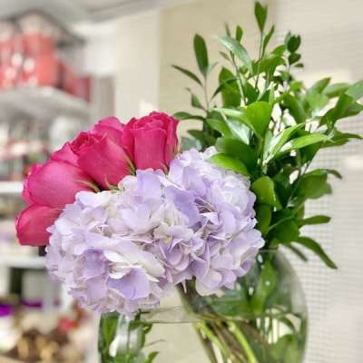 Round glass vase with pink roses, pale lavender hydrangeas, and tall green foliage.