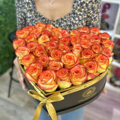 Heart-shaped black box filled with orange and yellow roses and tied with a gold ribbon