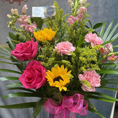 Bouquet of pink roses, sunflowers, and carnations in a purple vase with a pink ribbon