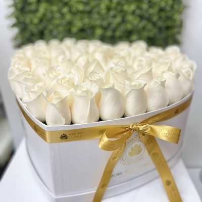 Heart-shaped white box filled with tightly packed ivory roses and tied with a gold satin ribbon.