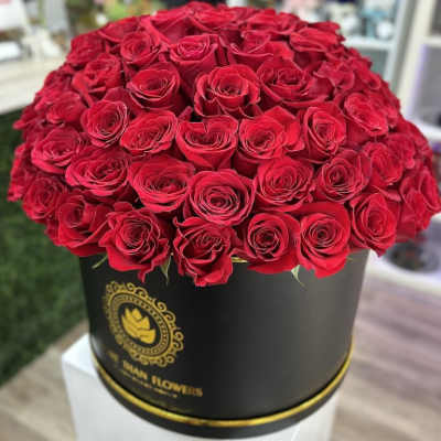 Round black hatbox filled with many red roses on a white pedestal.