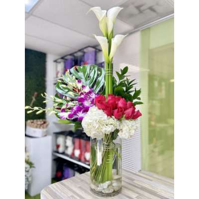 Tall modern arrangement with calla lilies, orchids, roses and hydrangeas in a glass vase with a monstera leaf