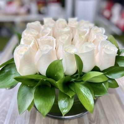 Low arrangement of cream roses tightly packed in a round glass vase with a collar of glossy green leaves.