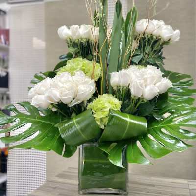 Tall modern arrangement of white roses and green hydrangeas in a square glass vase with large tropical leaves.