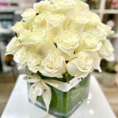Compact arrangement of cream roses in a clear square glass vase with a ribbon bow.