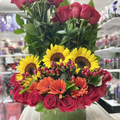 Tall arrangement of red roses, yellow sunflowers, and orange gerbera daisies in a round glass vase.