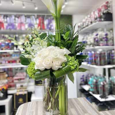 Tall white calla lilies and white roses with green accents arranged in a clear glass cylinder vase.