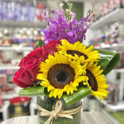 Mixed arrangement of sunflowers, red roses, and purple orchids in a glass cylinder vase with raffia bow