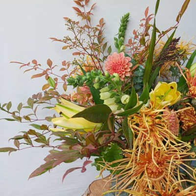 Asymmetrical arrangement with peach dahlias, yellow lilies and orange protea in a vase against a light background
